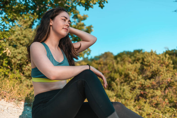 Young sporty woman resting, sitting on the grass. Forest in the background. Sports, fitness and healthy lifestyle tracking concept. - Φωτογραφία, εικόνα