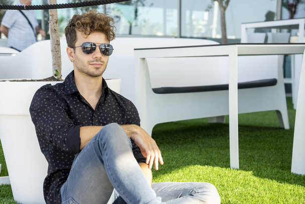 A young man wearing sunglasses and a black shirt and sitting on a chair outdoors - Photo, image