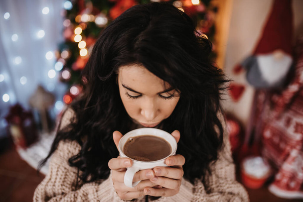 A Caucasian female holding cup of coffee with colorful Christmas tree on background at Christmas Eve - Fotó, kép