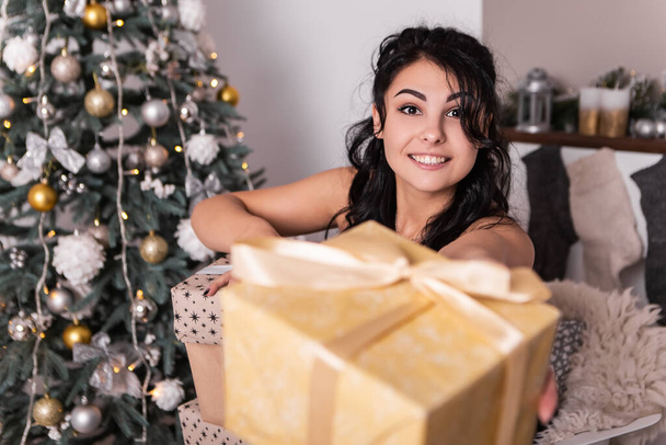 Brunette smiling happy woman opening wrapped ribbon gift box present, New Year evening, Christmas decorated tree. Lifestyle and shopping concept. Biracial girl greeting congratulating with holiday - Foto, immagini