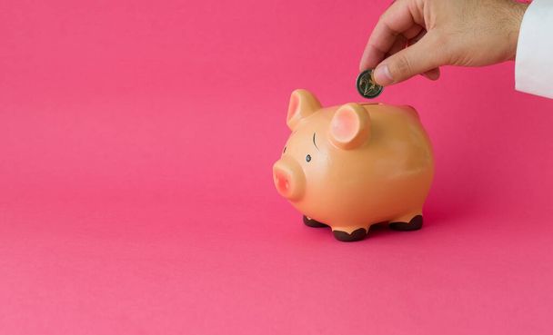 A man putting a coin into a piggy bank on pink background to save money - Foto, Imagem