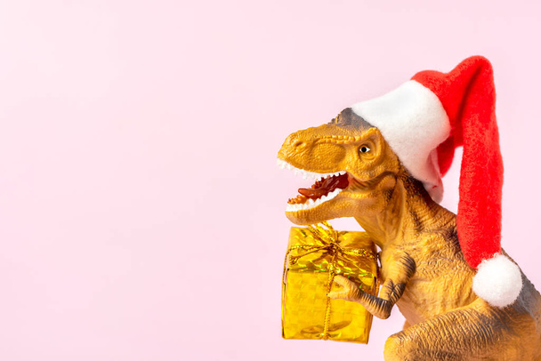 Dinosauro Rex in rosso cappello di Babbo Natale tiene scatola regalo d'oro nelle sue zampe su sfondo rosa Capodanno o la vigilia di Natale Art holiday card Idea creativa per il concetto di Buon Natale. - Foto, immagini