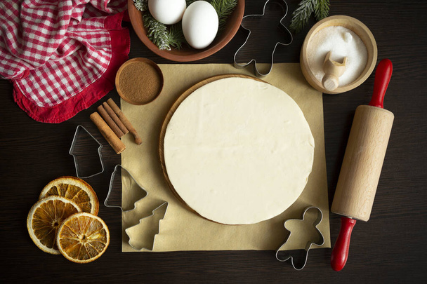 Culinary Christmas background with baking ingredients - round sheet of dough on the podium, cookie cutters, rolling pin, sugar, eggs, dried orange, two kinds of cinnamon, parchment, apron. Flat lay - Photo, Image