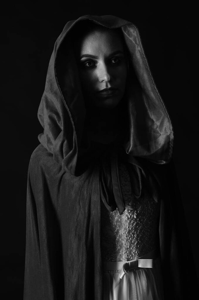 mooie vrouw gekleed in jurk en mantel poseren op zwarte achtergrond, zwart en wit foto - Foto, afbeelding