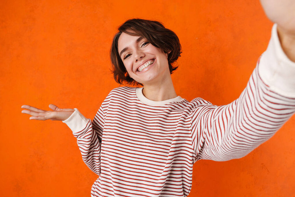 Young brunette woman holding copyspace while taking selfie photo isolated over orange wall - Fotografie, Obrázek
