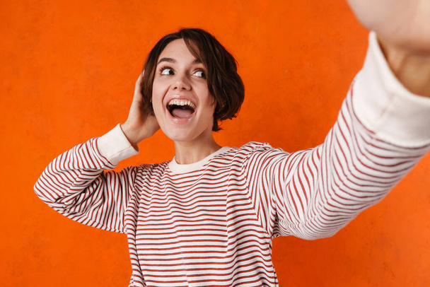 Young brunette woman laughing while taking selfie photo isolated over orange wall - Foto, imagen