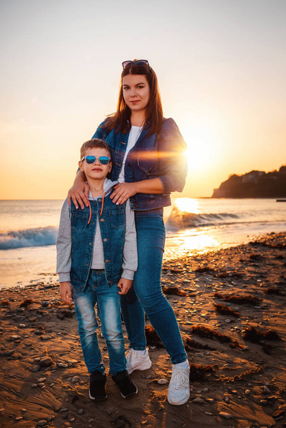 若い笑顔の母親と彼女の息子がビーチに立っている。背景には海と夕日。幸せな子供時代と母親の概念. - 写真・画像