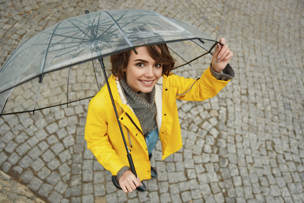Caucasian girl in yellow raincoat with transparent umbrella in city. Young beautiful woman wearing rain outfit while walking on street at daytime. Concept of modern woman lifestyle at autumn season.  - Foto, Imagen