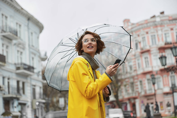 Caucasian girl in yellow raincoat with transparent umbrella in city. Young beautiful woman wearing rain outfit while walking on street at daytime. Concept of modern woman lifestyle at autumn season.  - Foto, imagen