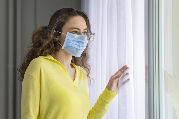 Young woman in isolation at home near window. Woman in quarantine looking through the window. Covid-19 virus protection. - Foto, imagen