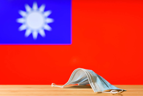 Una maschera medica si trova sul tavolo sullo sfondo della bandiera di Taiwan. Il concetto di un regime di maschera obbligatoria per i residenti del paese e turisti a Taiwan durante una pandemia. - Foto, immagini