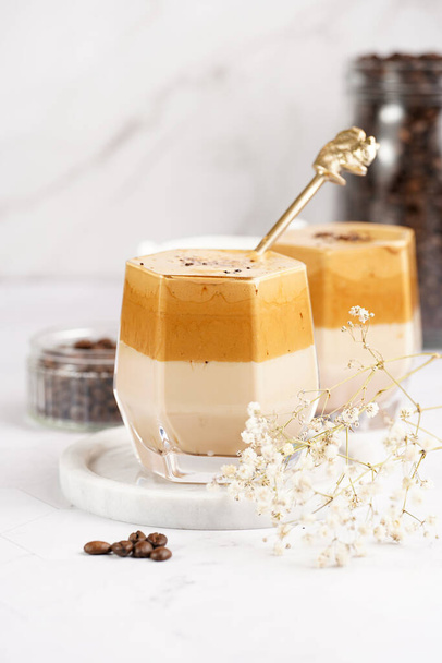 Two glasses with trendy korean beverage dalgona coffee latte - whipped instant coffee, sugar and hot water and milk layers on a marble board, white surface - Φωτογραφία, εικόνα