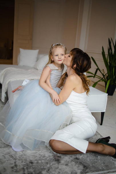 Beauty mother and little daughter in dress hugging and sitting on the bed home. - Photo, Image