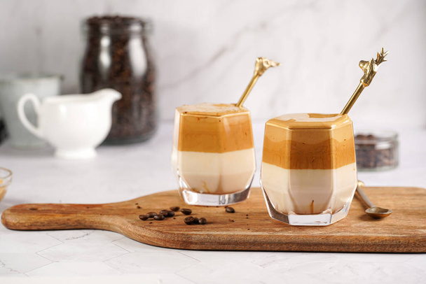 Δύο ποτήρια με μοντέρνο κορεάτικο ρόφημα dalgona καφέ latte - στιγμιαίος σαντιγί καφές, ζάχαρη και ζεστό νερό και στρώματα γάλακτος σε ξύλινη σανίδα, λευκή επιφάνεια - Φωτογραφία, εικόνα