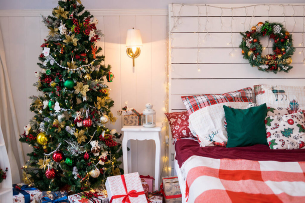  Kerstvakantie met boom en geschenken. Ruime witte lichte slaapkamer in een loft stijl met een versierde kerstsparren, slinger, bed met kussens. Binnenkamer met kerstmis. Hoge kwaliteit foto - Foto, afbeelding