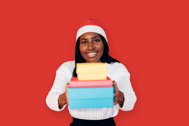όμορφο κορίτσι της αφρικανικής εθνικότητας φορώντας ένα χριστουγεννιάτικο καπέλο μοιράζει μερικά δώρα - Φωτογραφία, εικόνα
