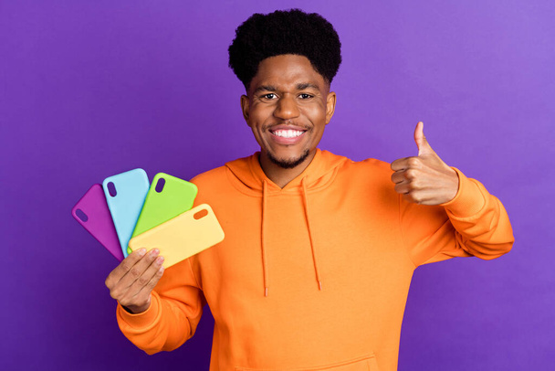 Photo of brunette afro american man make thumb up hold phone case wear hoodie orange sweater isolated on purple color background - Foto, Bild