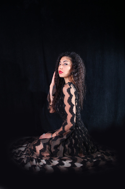 黒い背景の上に座って網タイツと髪の巻き毛のドレスの女性  - 写真・画像