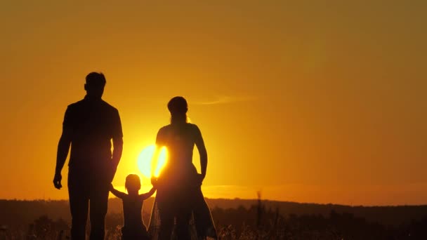 Šťastná rodina a dítě se procházejí v létě při západu slunce v parku. Týmová práce. Rodiče a dcera chodí spolu a drží se za ruce na poli na slunci. Silueta šťastné rodiny, jak spolu cestují ven - Záběry, video