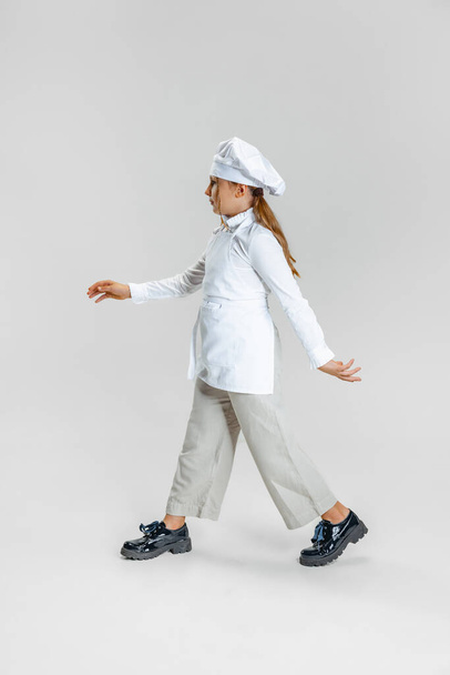 Little Caucasian girl in white cook uniform and huge chefs hat going isolated on white studio background. - Foto, immagini
