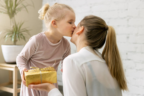 pieni tytär antaa lahjan ja suudella äitiään kotona. syntymäpäivä, naisten päivä tai äitien päivä käsite - Valokuva, kuva