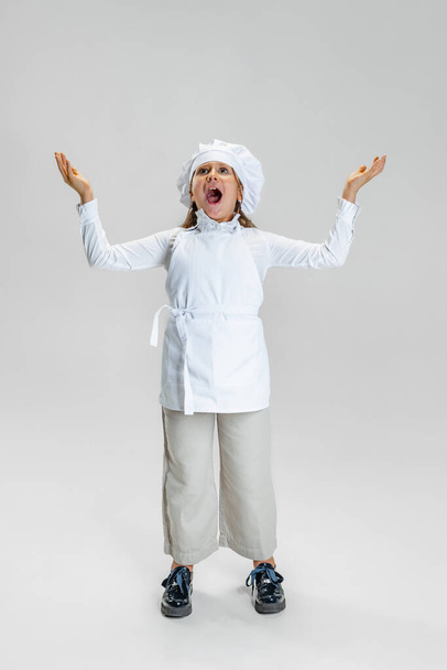 Portrait of little cute girl in white cook uniform and huge chefs hat shouting isolated on white studio background. - Zdjęcie, obraz