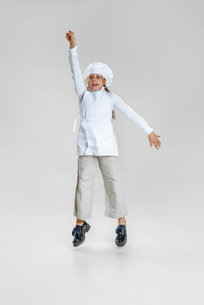 Dynamic image of little cute girl in white cook uniform and huge chefs hat jumping isolated on white studio background. - Φωτογραφία, εικόνα