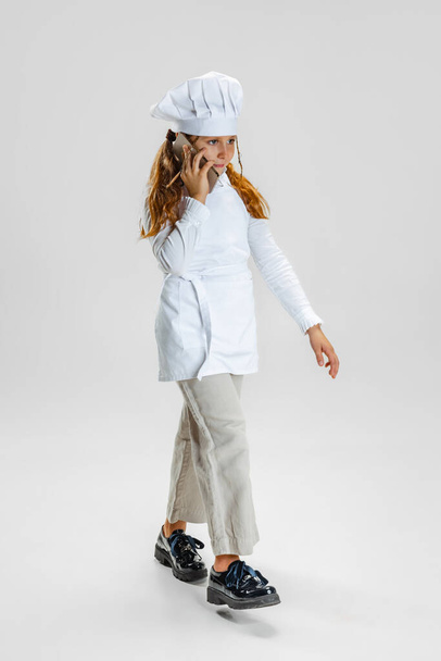 Full-length portrait of little girl in white cook uniform and huge chefs hat posing isolated on white studio background. - Foto, immagini