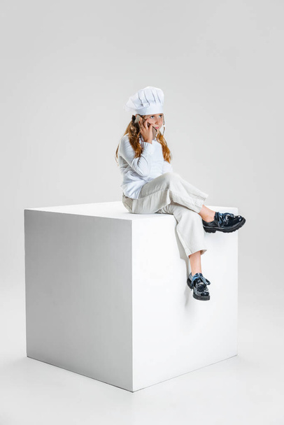 Photography of little girl, student in white cook uniform and huge chefs hat sitting on huge box isolated on white studio background. - Foto, imagen