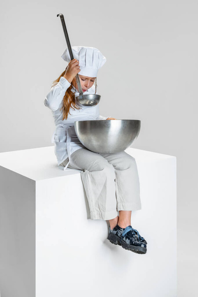 Fotografía de niña, estudiante en uniforme de cocinero blanco y sombrero de chefs enormes sentados en una caja enorme aislada sobre fondo de estudio blanco. - Foto, imagen