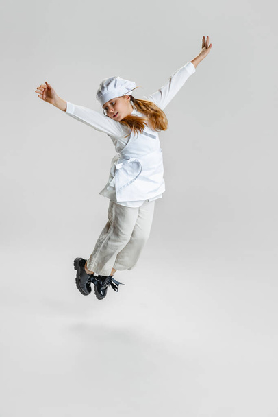Dynamic image of little cute girl in white cook uniform and huge chefs hat flying isolated on white studio background. - Fotografie, Obrázek