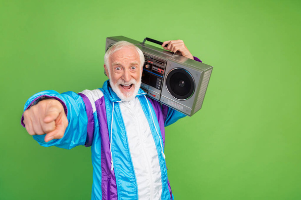 Photo of sweet excited mature guy dressed blue sport suit smiling pointing finger you listening boom box isolated green color background - Foto, imagen