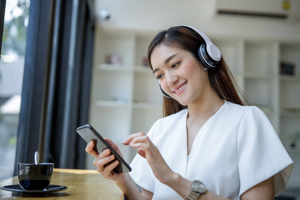  asiatique femme d'affaires assis près de la fenêtre en verre et en streaming écouter de la musique du téléphone mobile au casque sans fil et boire du café heureux   - Photo, image
