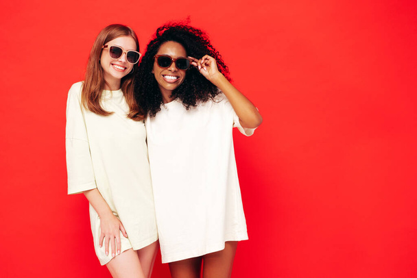 Two young beautiful smiling international hipster female in trendy summer clothes. Sexy carefree women posing near red wall in studio. Positive models having fun. Concept of friendship. In sunglasses - Zdjęcie, obraz