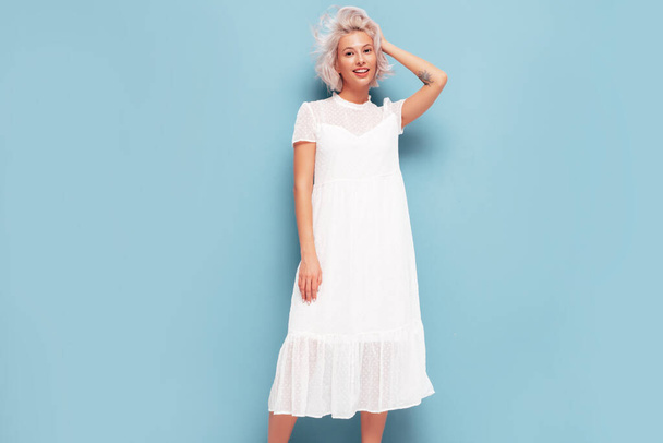 Portrait of young beautiful smiling female in trendy summer white dress. Sexy carefree blond woman posing near blue wall in studio. Positive model having fun indoors. Cheerful and happy - Foto, imagen