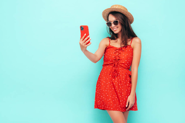 Young beautiful smiling female in trendy summer red dress. Sexy carefree woman posing near blue wall in studio. Positive brunette model having fun. In sunglasses and hat. Takes selfie - Foto, afbeelding