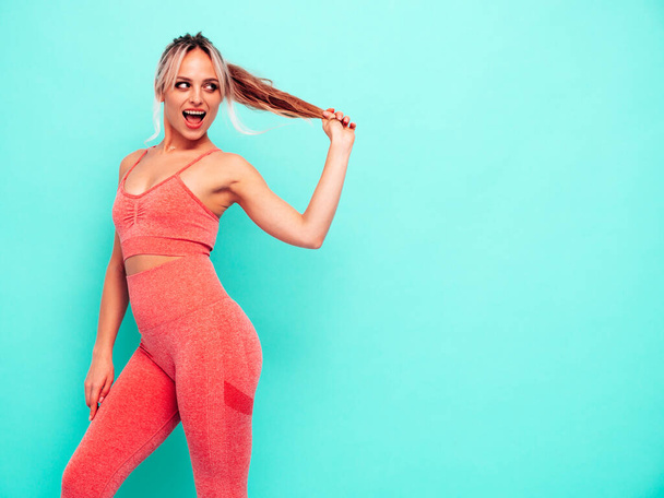 Portrait of fitness smiling woman in pink sports clothing. Young beautiful model with perfect body.Female posing near blue wall in studio.Cheerful and happy. Holding her hair tail - Φωτογραφία, εικόνα