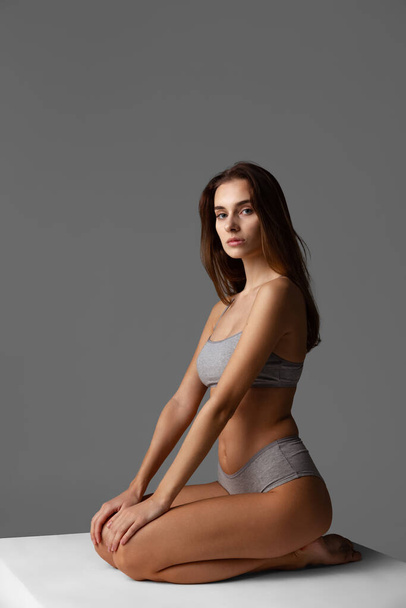 Studio shot of young beautiful slim girl in grey underwear sitting on box isolated over gray studio background. Natural beauty concept. - Fotó, kép