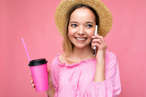 Portrait of beautiful positive emotional young blonde woman wearing pink crop blouse and straw hat talking on smartphone while drinking beverage isolated on pink background with copy space. Phone - Photo, Image