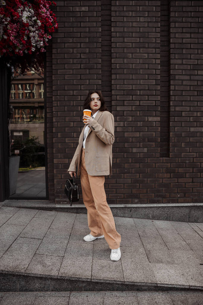 Tall stylish girl with cofee in a beige oversized jacket and orange pants on brick wall background - Photo, Image