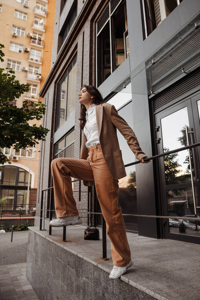  Ψηλό κομψό κορίτσι με υπερμεγέθης σακάκι ποζάρουν κοντά στο κιγκλίδωμα και βήματα στο φόντο της πόλης - Φωτογραφία, εικόνα