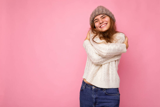 Foto de hermosa mujer morena sonriente feliz aislada sobre la pared de fondo rosa con suéter blanco de punto y sombrero cálido gris y divertirse - Foto, imagen