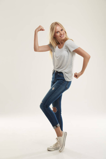 Young joking european girl posing like bodybuilder and looking at camera. Beautiful blonde female teenager with blue eyes wear white t-shirt and jeans. Isolated on white background. Studio shoot - Zdjęcie, obraz