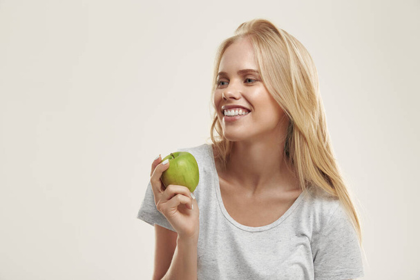 Młoda, zadowolona Europejka trzymająca zielone jabłko. Piękna blondynka z niebieskimi oczami nosi białą koszulkę. Odizolowany na białym tle. Zdjęcia studyjne - Zdjęcie, obraz