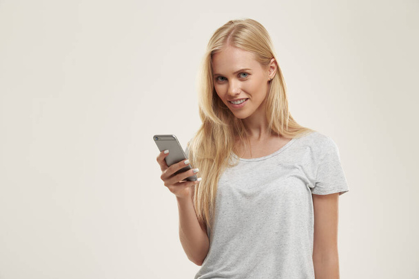 Jong lachend Europees meisje met een mobiele telefoon. Mooie blonde vrouwelijke tiener met blauwe ogen dragen witte t-shirt en kijken naar de camera. Geïsoleerd op witte achtergrond. Studio shoot. Kopieerruimte - Foto, afbeelding