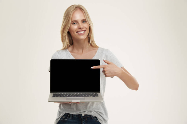Young smiling european girl showing laptop computer and looking at camera. Beautiful blonde female teenager with blue eyes wear white t-shirt. Isolated on white background. Studio shoot - Fotó, kép