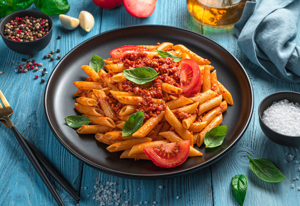 Penne pasta met bolognese saus, met verse tomaten en basilicum op een blauwe achtergrond. zijaanzicht, close-up. - Foto, afbeelding