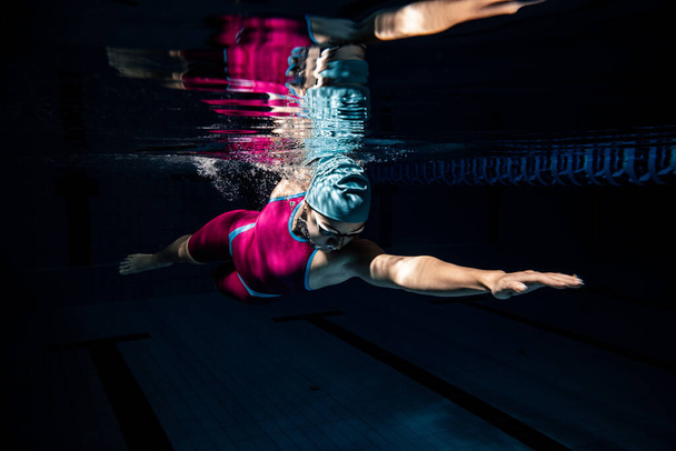 One female swimmer in swimming cap and goggles training at pool, indoors. Underwater view of swimming movements details. - Photo, image