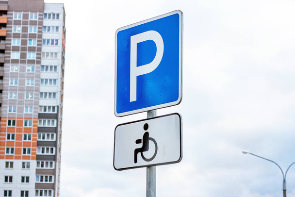 Niebiesko-biały znak parkingowy z wózkiem inwalidzkim dla osób niepełnosprawnych na tle nieba i budynków miejskich. - Zdjęcie, obraz