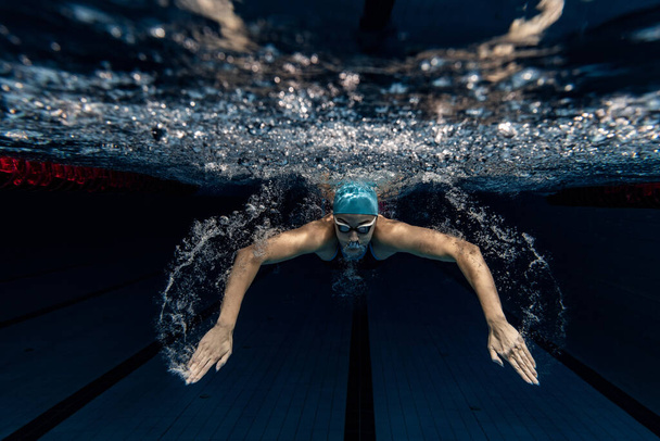 Professional female swimmer in swimming cap and goggles in motion and action during training at pool, indoors. Underwater shooting - Foto, afbeelding